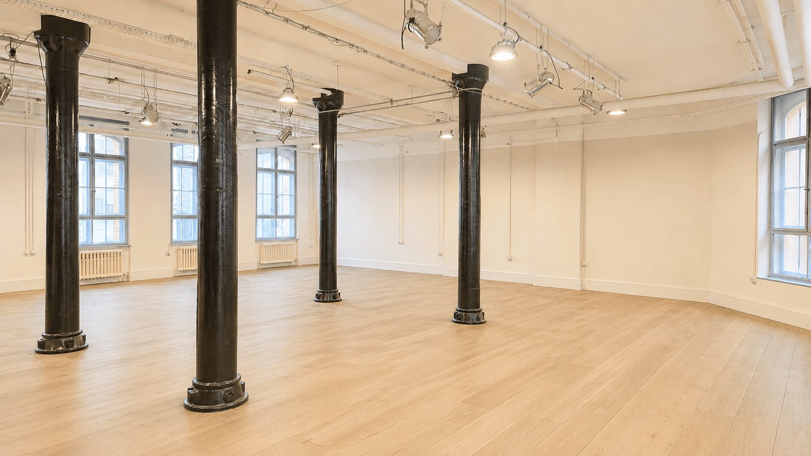 Empty industrial-style loft studio with four black support columns, light wood floor, and large arched windows letting in natural light