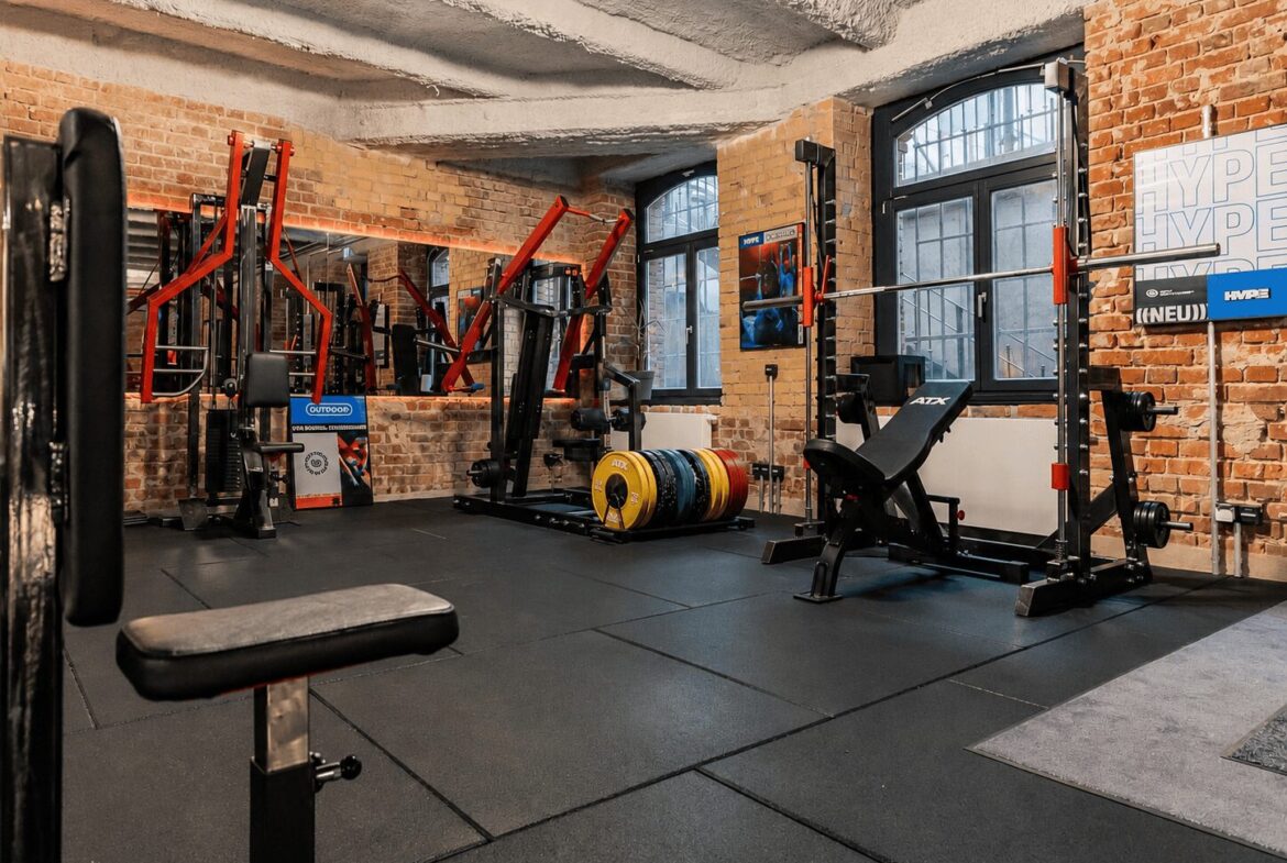 A brick-walled gym with weight benches, racks, and a barbell setup near large windows. colorful plates stacked nearby.