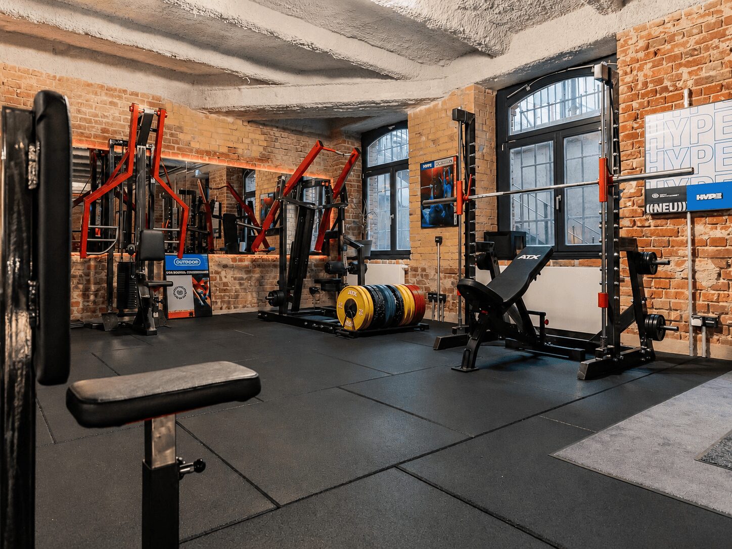 A brick-walled gym with weight benches, racks, and a barbell setup near large windows. colorful plates stacked nearby.