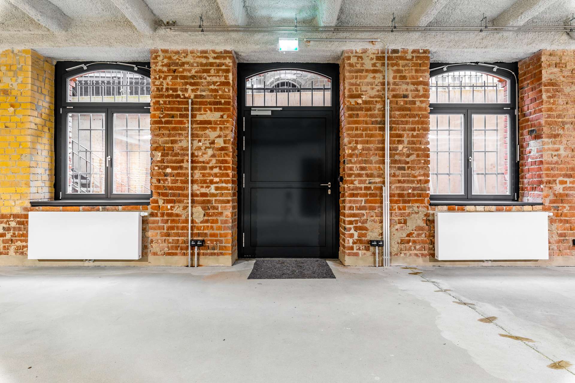 Industrial brick interior with a black central door and arched windows on each side, metal window bars, and visible radiators below.