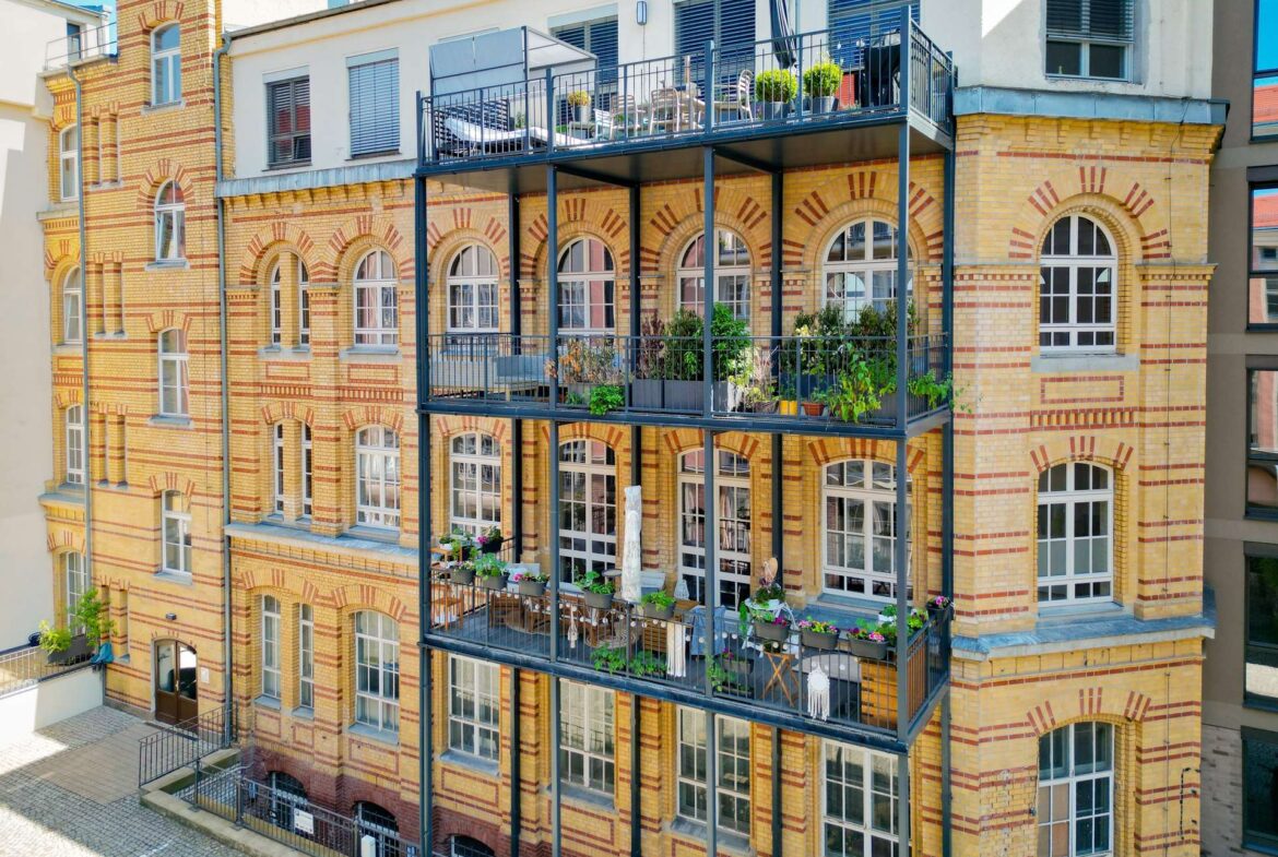 Brick building with arched windows and stacked black metal balconies filled with potted plants on each level.