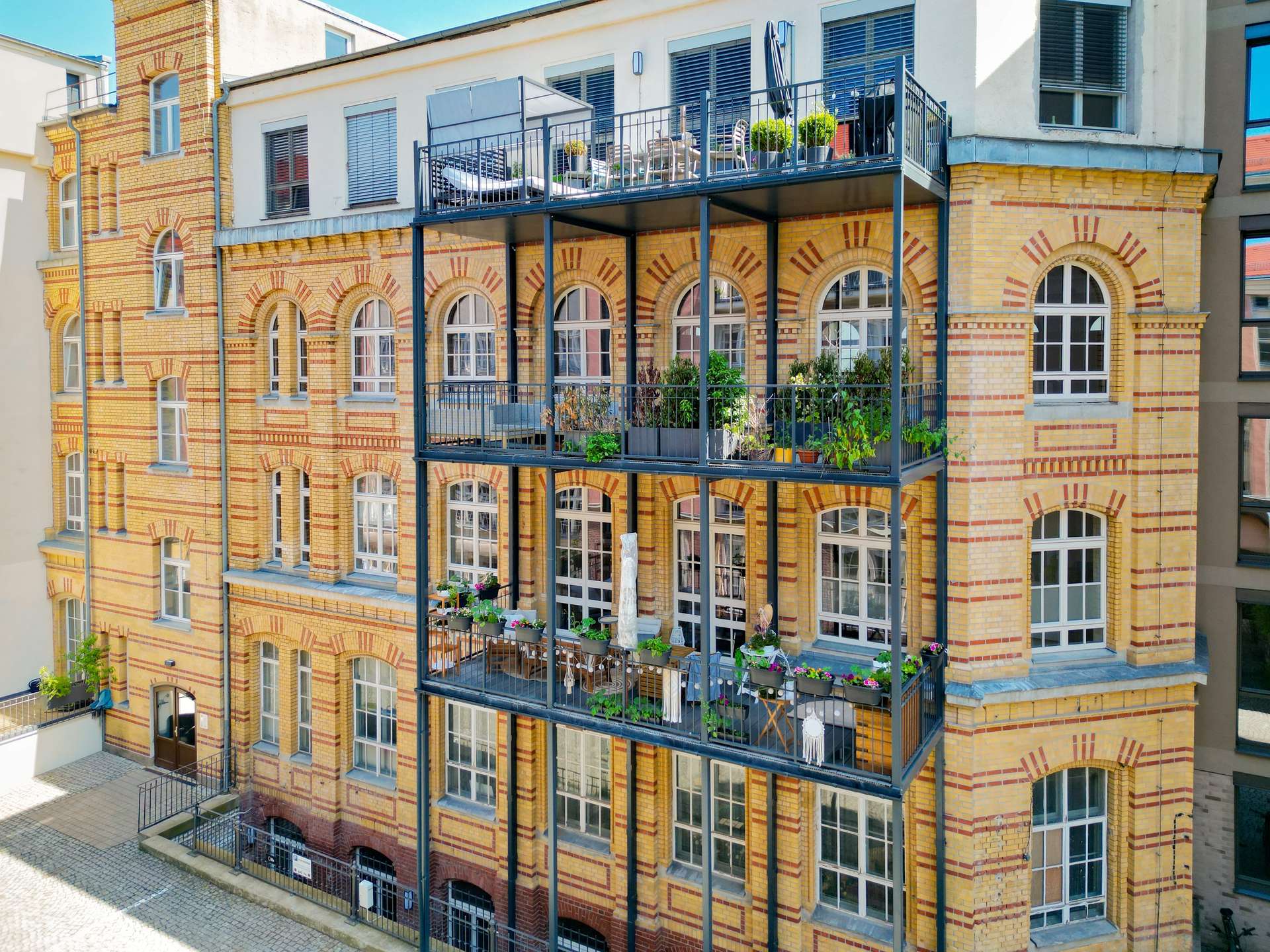Brick building with arched windows and stacked black metal balconies filled with potted plants on each level.