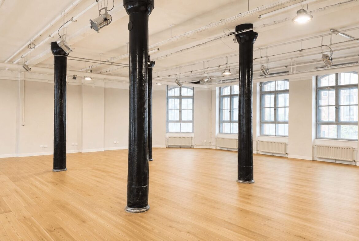 Empty industrial-style studio with a hardwood floor, white walls, large windows, and several black support columns.