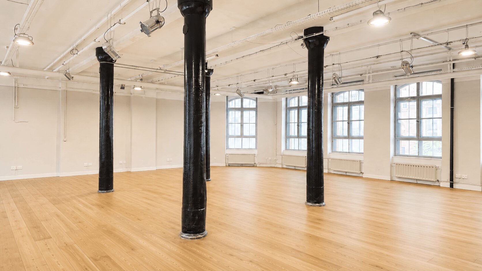 Empty industrial-style studio with a hardwood floor, white walls, large windows, and several black support columns.