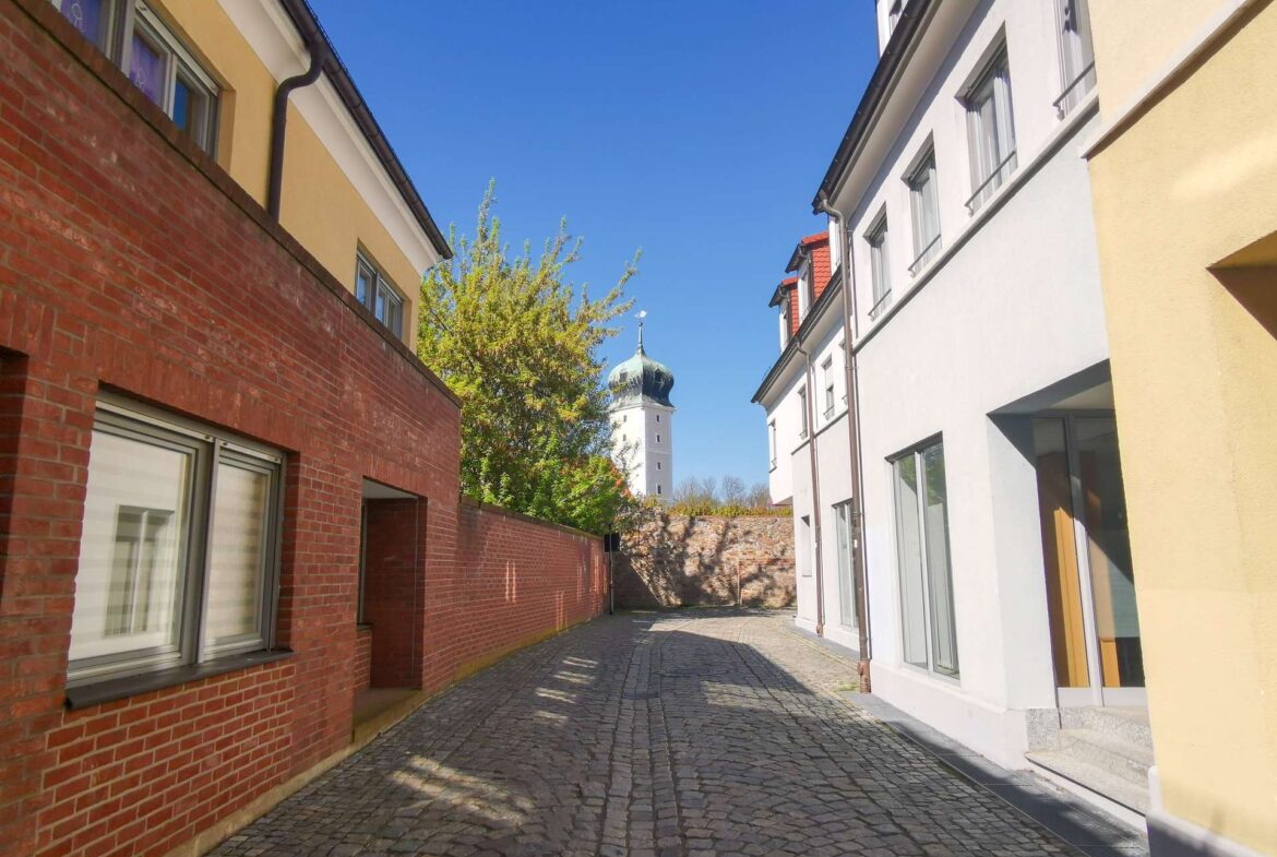 Narrow cobblestone alley between a red brick wall on the left and a pale, white-yellow building on the right, with a church tower in the distance under a blue sky.