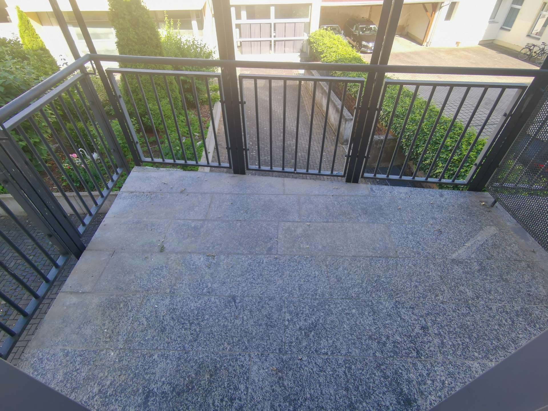 View from a balcony looking down onto a narrow brick pathway flanked by green hedges and a metal railing enclosure in the foreground.