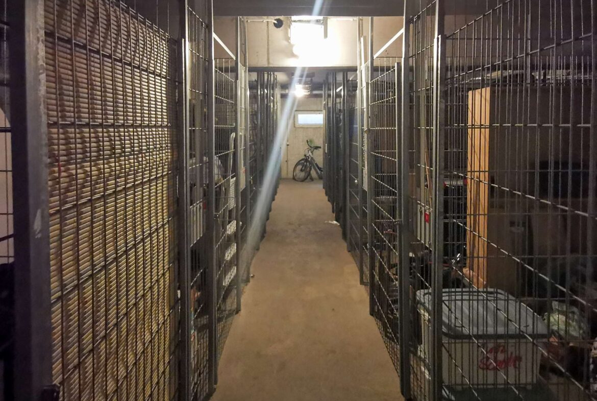 Narrow storage corridor with metal cages on both sides; a bicycle parked at the far end beneath bright overhead lights.