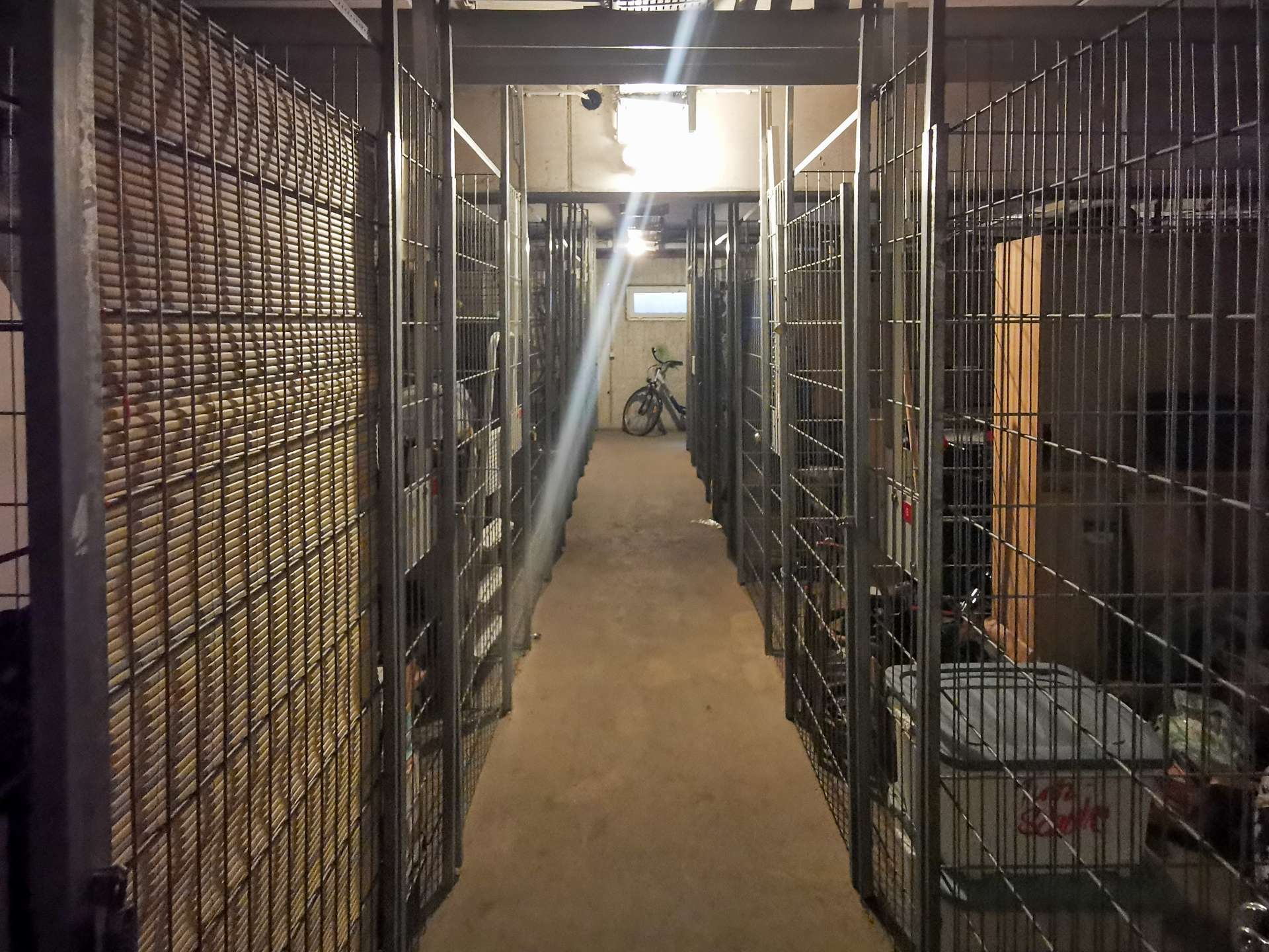 Narrow storage corridor with metal cages on both sides; a bicycle parked at the far end beneath bright overhead lights.