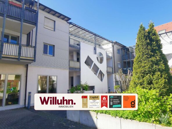Modern apartment building with glass-walled walkways and a large Willuhn Immobilien sign featuring awards on the front.