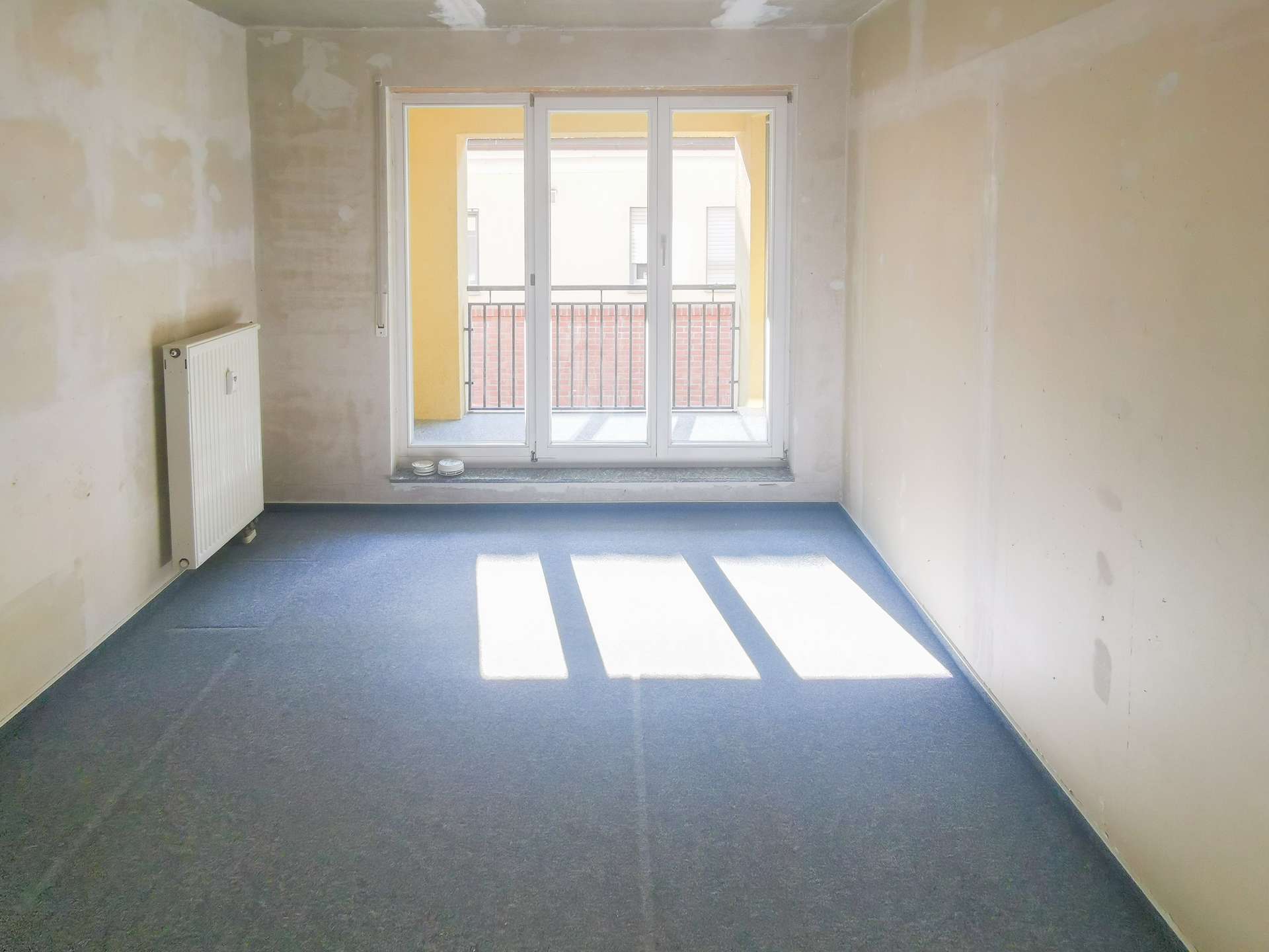 Empty unfinished room with a radiator on the left and a large glass door to a balcony, sunlight streaming in from outside on a blue floor.