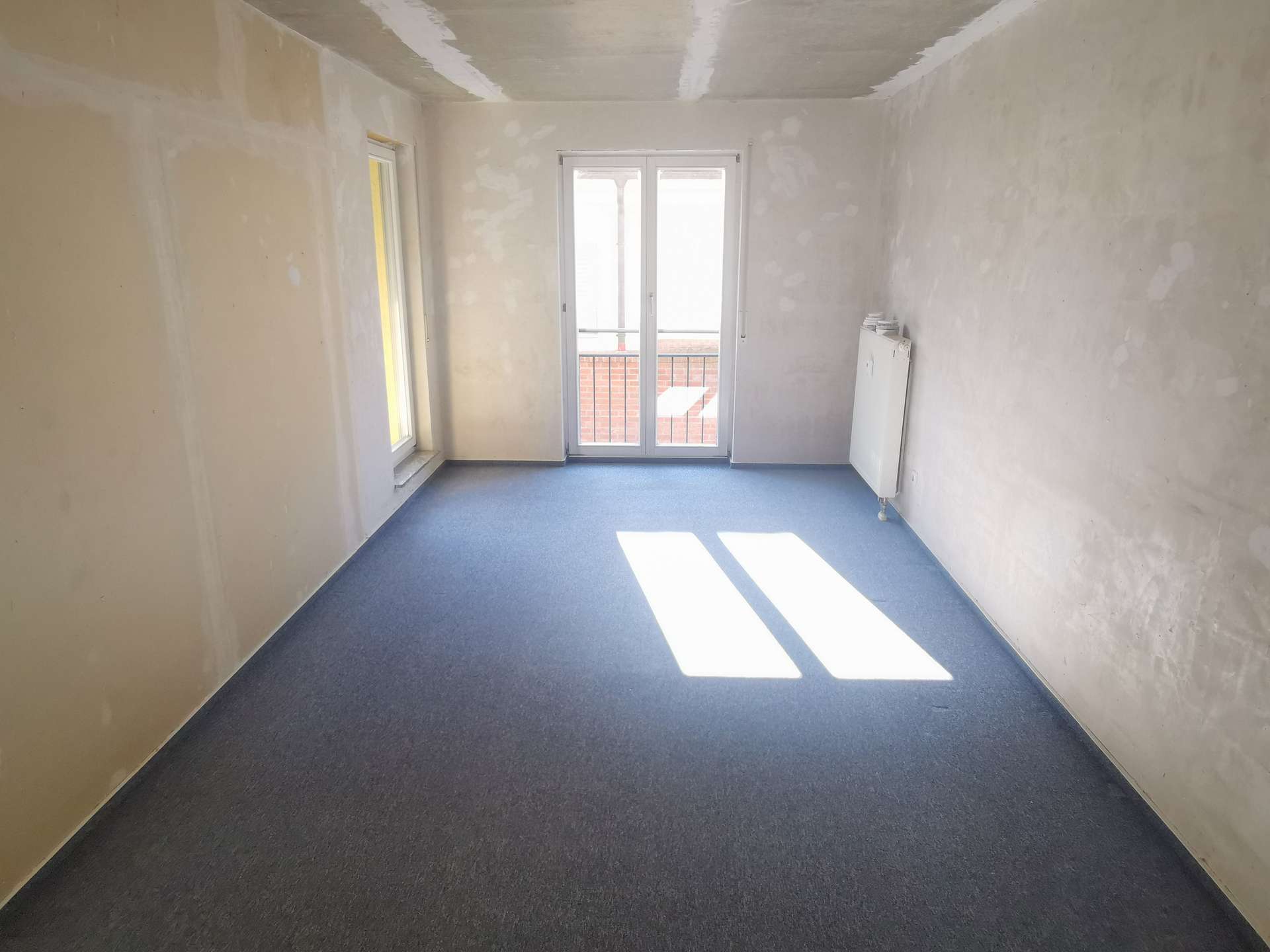 Unfinished rectangular room with patchy white walls, blue carpet, a white radiator on the right, and a glass door to a balcony letting in sunlight.