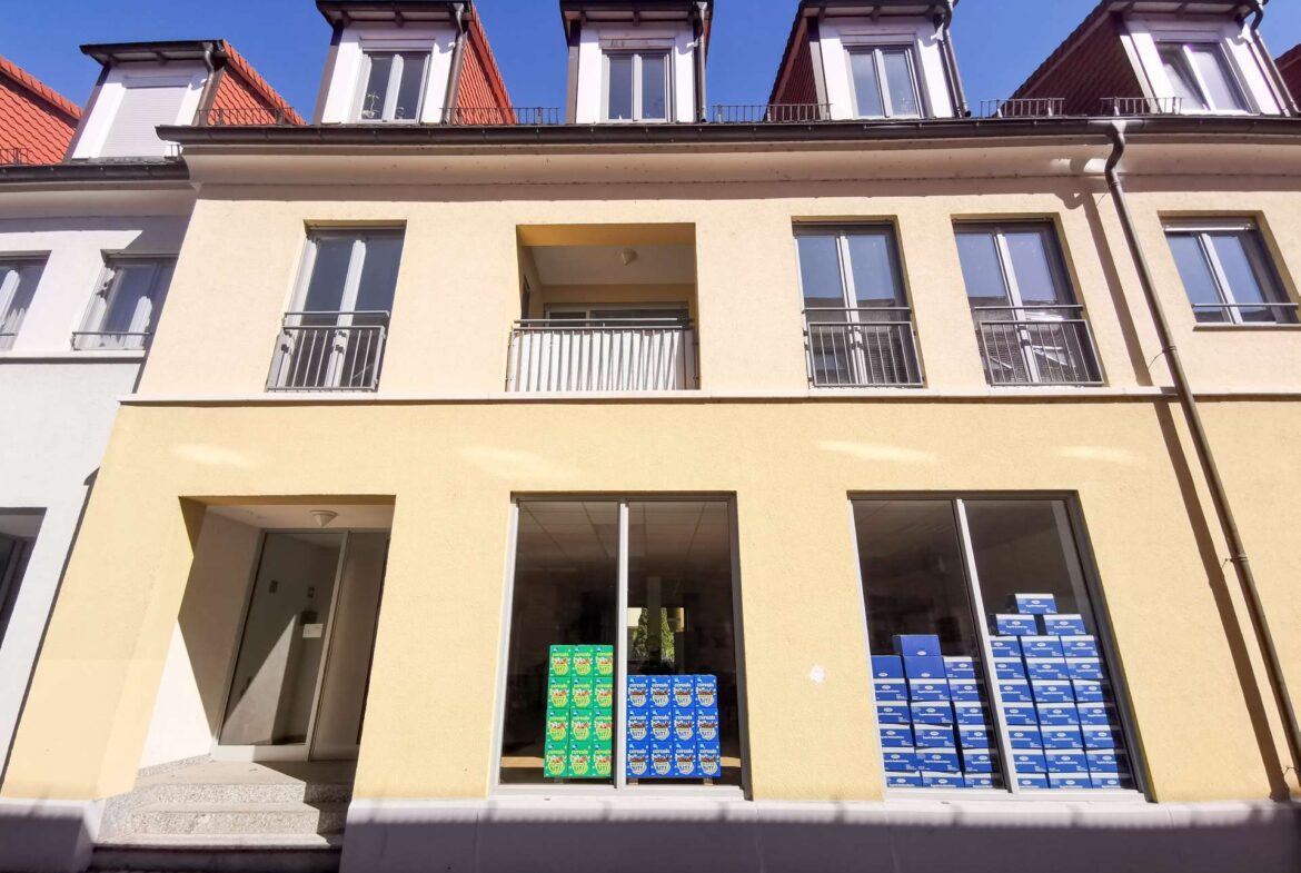 Ground-floor store with large windows displaying stacks of blue and green product boxes against a yellow facade and beige entryway left of center