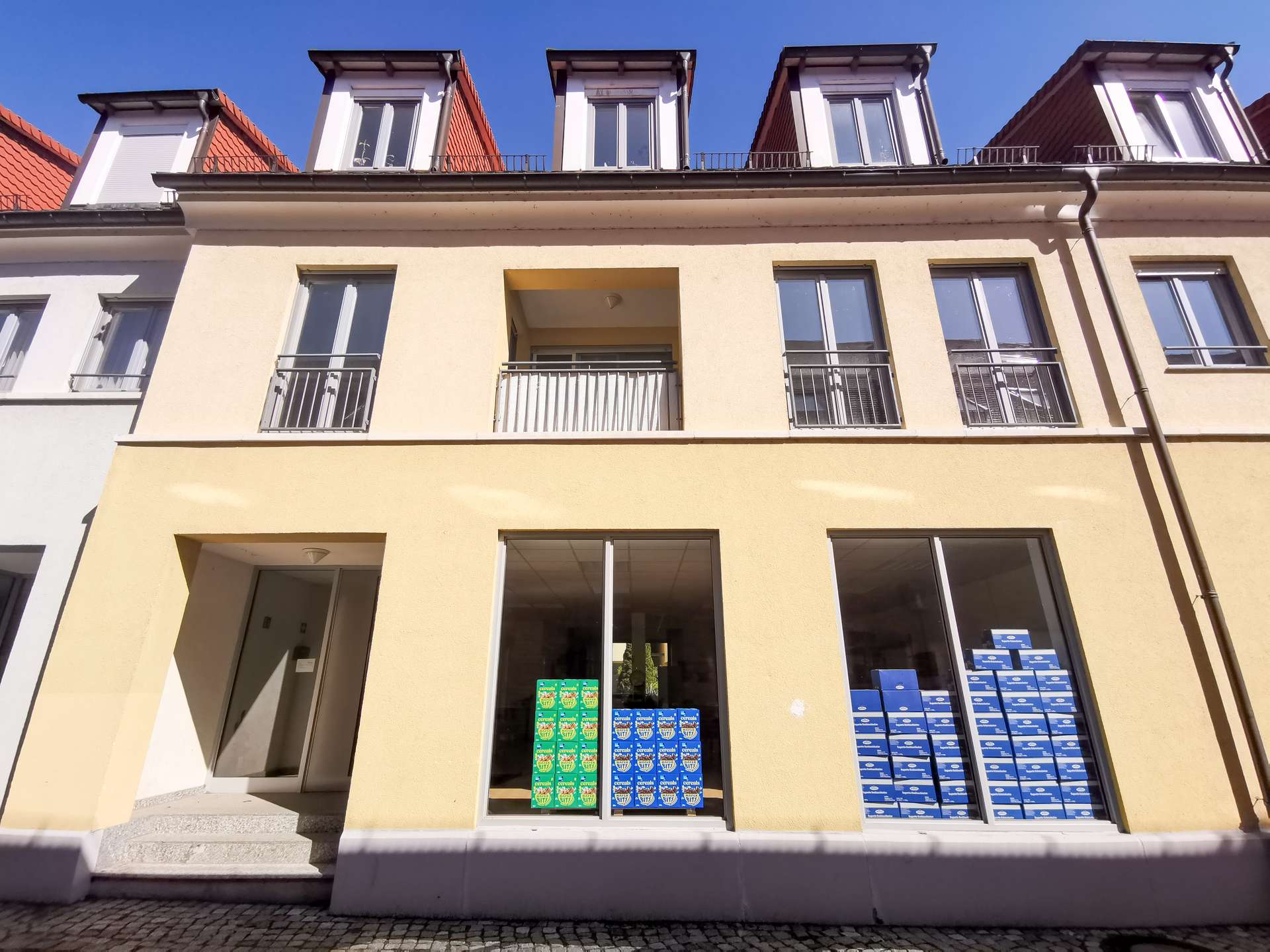 Ground-floor store with large windows displaying stacks of blue and green product boxes against a yellow facade and beige entryway left of center
