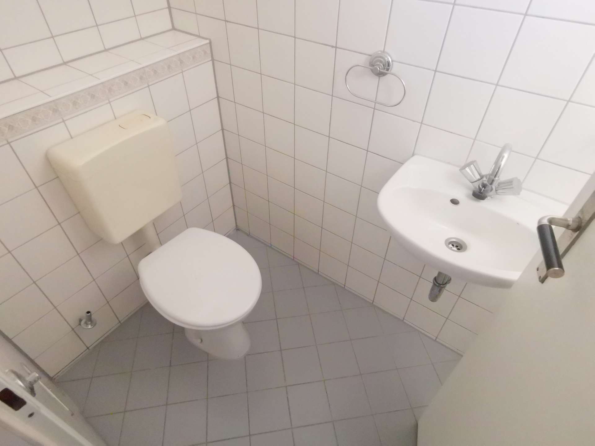 Compact white-tiled bathroom with a toilet on the left and a small wall-mounted sink with chrome faucet on the right, plus a chrome towel ring on the wall.