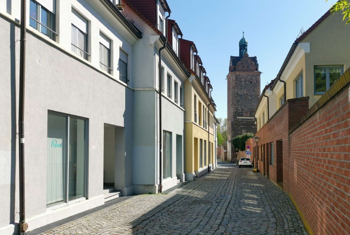 Narrow cobblestone street flanked by white and yellow townhouses, with a brick wall on the right and a distant historic tower under a blue sky.