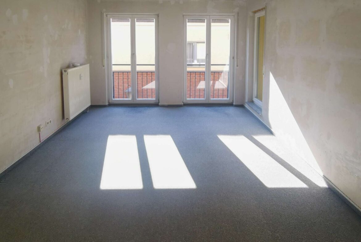Bright, unfinished room with three large glass doors leading to a balcony; sunlight casts rectangular shadows on the blue floor.