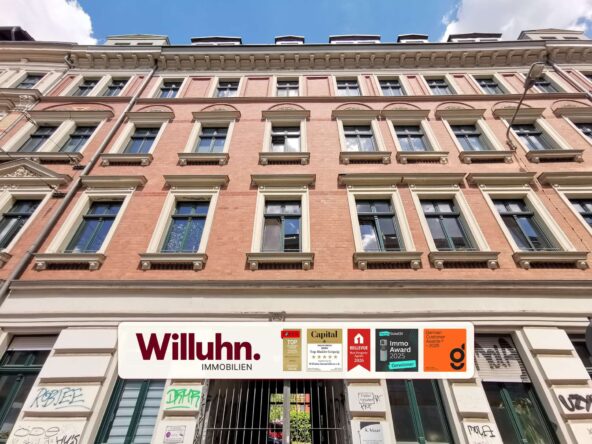 Facade of a pink brick building with many windows above a Willuhn Immobilien storefront sign and badges: real estate agency.