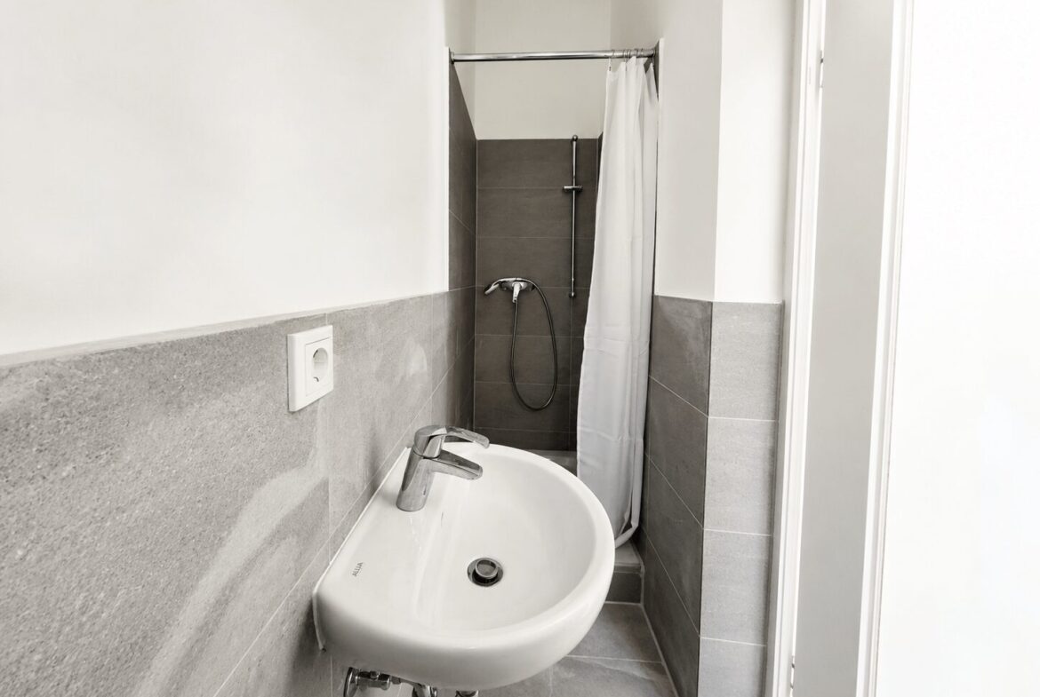 Small bathroom with a white wall-mounted sink, silver faucet, and gray tiled walls; a shower with a white curtain is in the back.