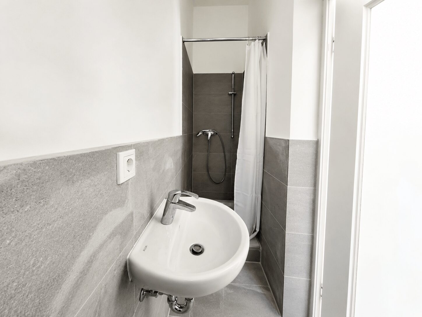 Small bathroom with a white wall-mounted sink, silver faucet, and gray tiled walls; a shower with a white curtain is in the back.