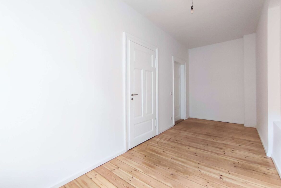 Empty white hallway with light wood floor, white doors along the left wall, and a bare hanging bulb overhead.