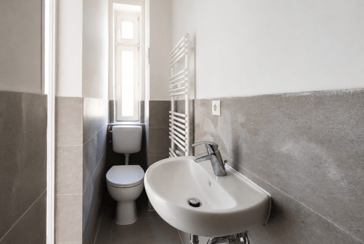 Small bathroom with a white toilet and pedestal sink, gray tile wainscoting, and a vertical white heated towel radiator by the wall; narrow window at the back letting in light.