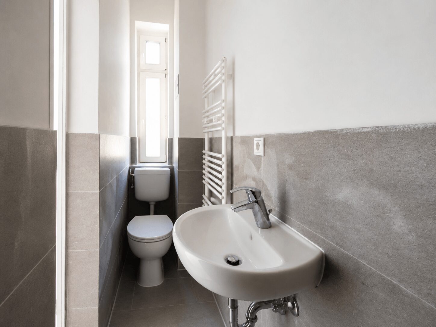 Small bathroom with a white toilet and pedestal sink, gray tile wainscoting, and a vertical white heated towel radiator by the wall; narrow window at the back letting in light.