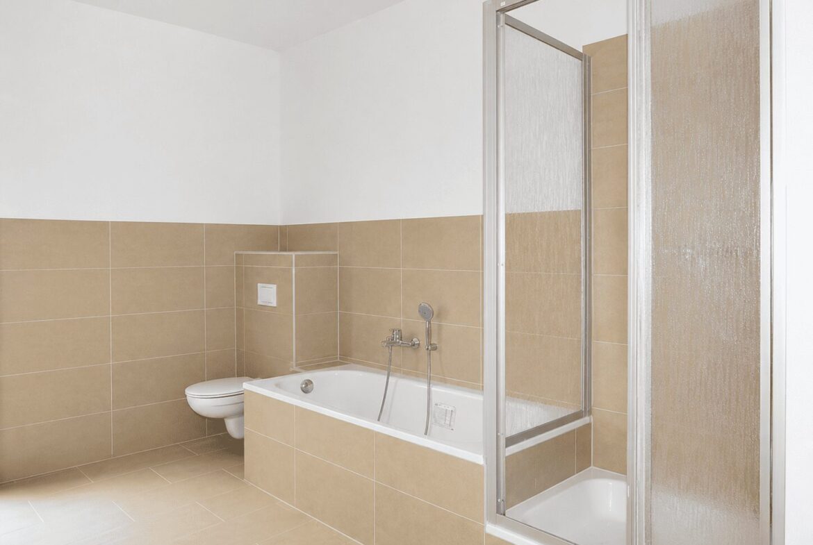 Bathroom with beige tile walls and floor, featuring a white built-in bathtub, wall-mounted toilet, and glass-front shower enclosure on the right.