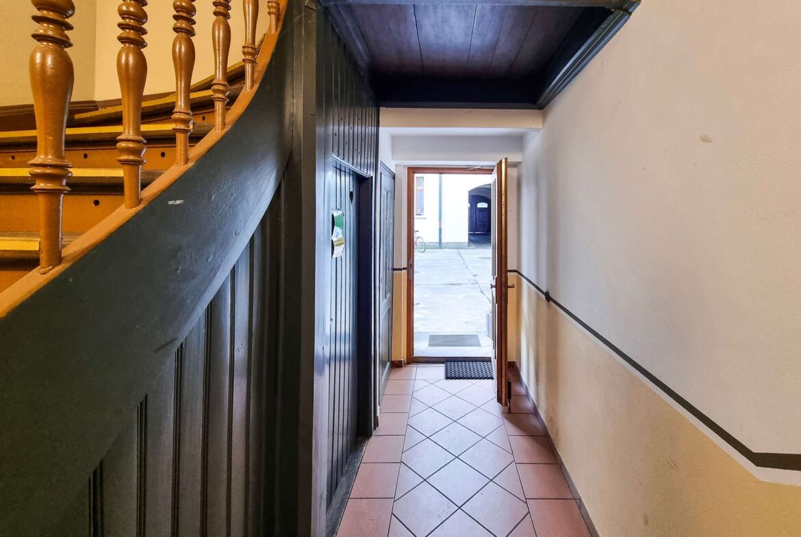 Narrow hallway with dark wooden paneling and a curved staircase on the left, open door at the far end showing a street outside and a bicycle.