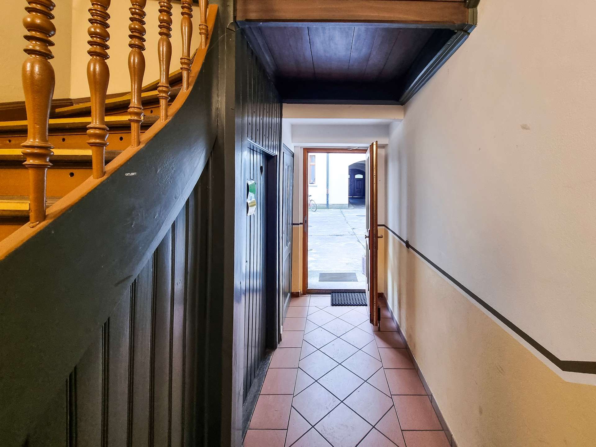 Narrow hallway with dark wooden paneling and a curved staircase on the left, open door at the far end showing a street outside and a bicycle.