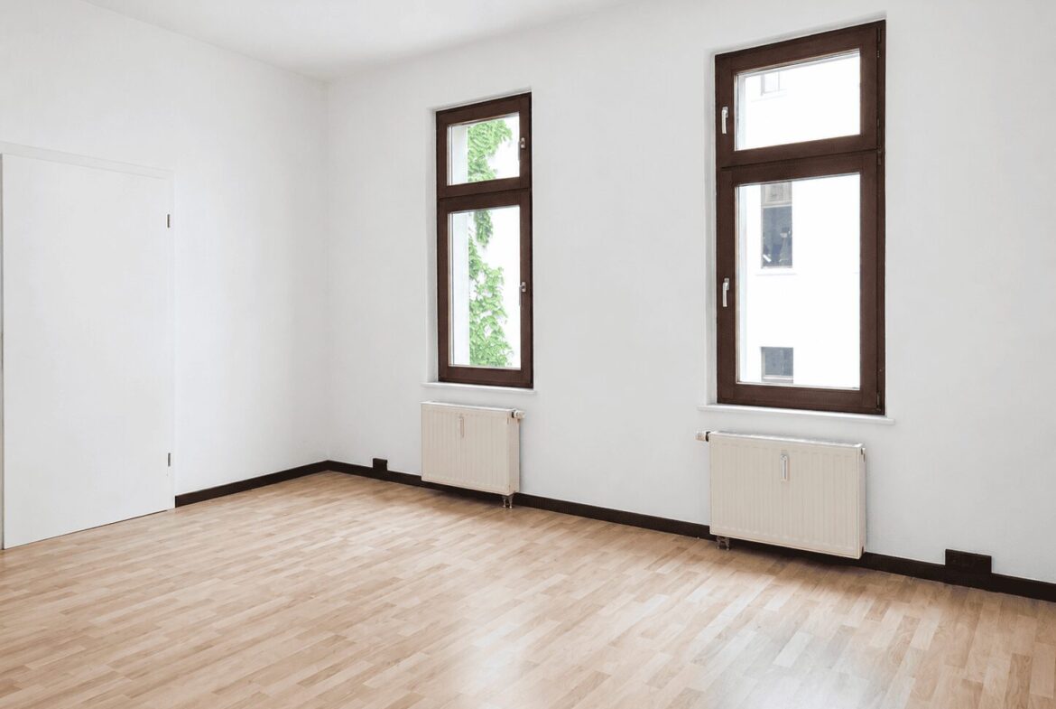 Bright empty room with light wood flooring, white walls, two tall brown-framed windows, and radiators beneath them.