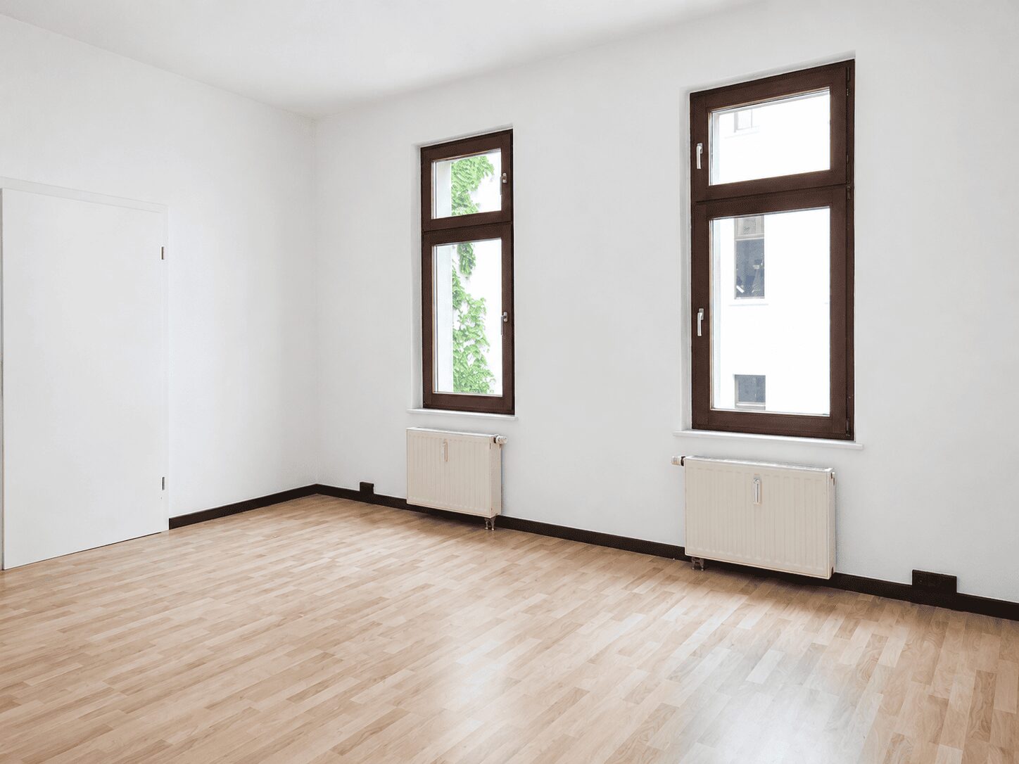 Bright empty room with light wood flooring, white walls, two tall brown-framed windows, and radiators beneath them.