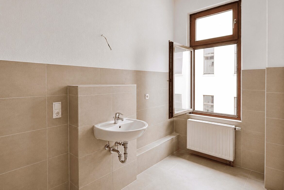Empty beige-tiled bathroom under renovation with a wall-mounted sink and exposed plumbing near a large window.