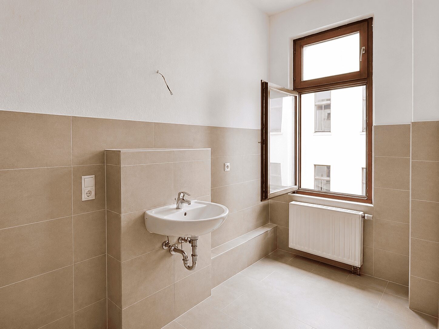 Empty beige-tiled bathroom under renovation with a wall-mounted sink and exposed plumbing near a large window.