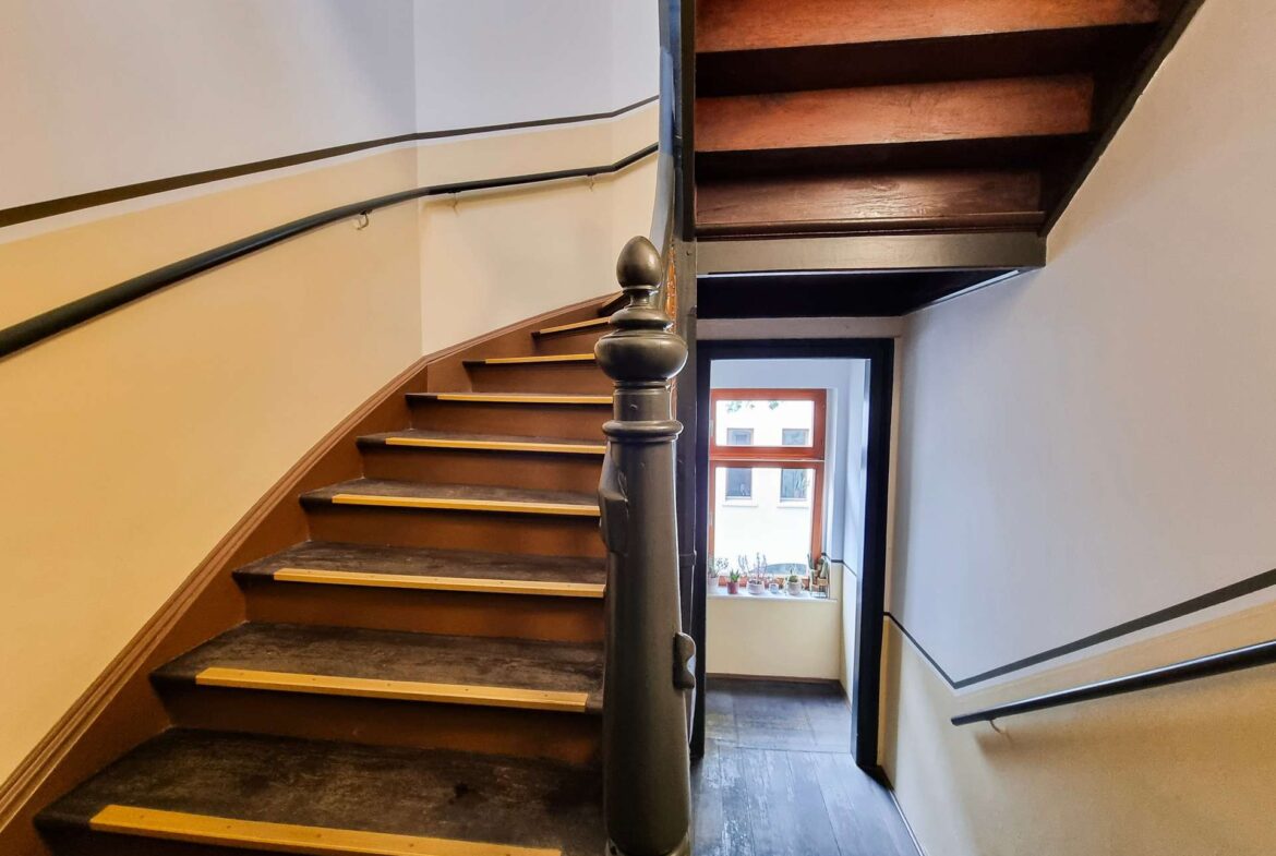 Wooden indoor staircase with a dark handrail, curving upward toward a doorway and window at the end.