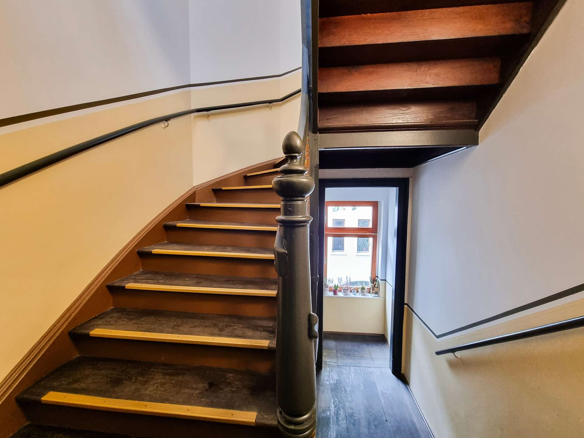 Wooden indoor staircase with a dark handrail, curving upward toward a doorway and window at the end.