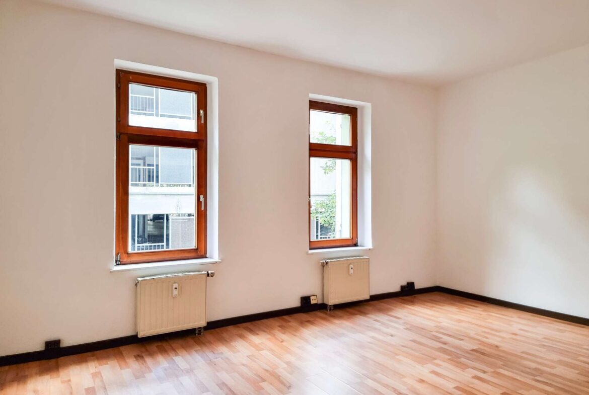 Empty room with two wooden-framed windows, radiators beneath, and light wood laminate flooring.