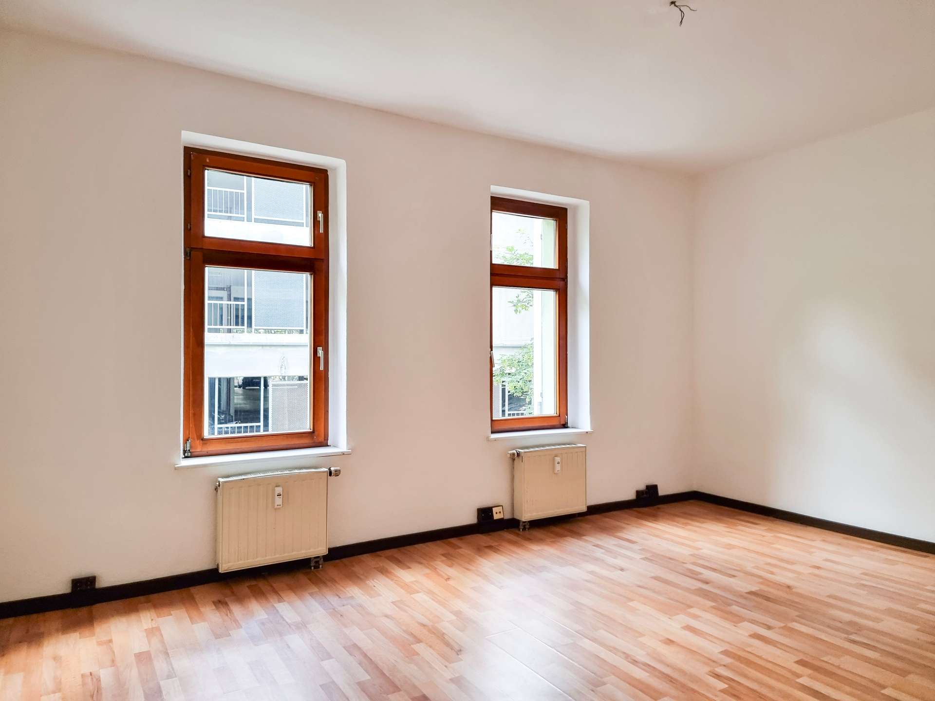 Empty room with two wooden-framed windows, radiators beneath, and light wood laminate flooring.
