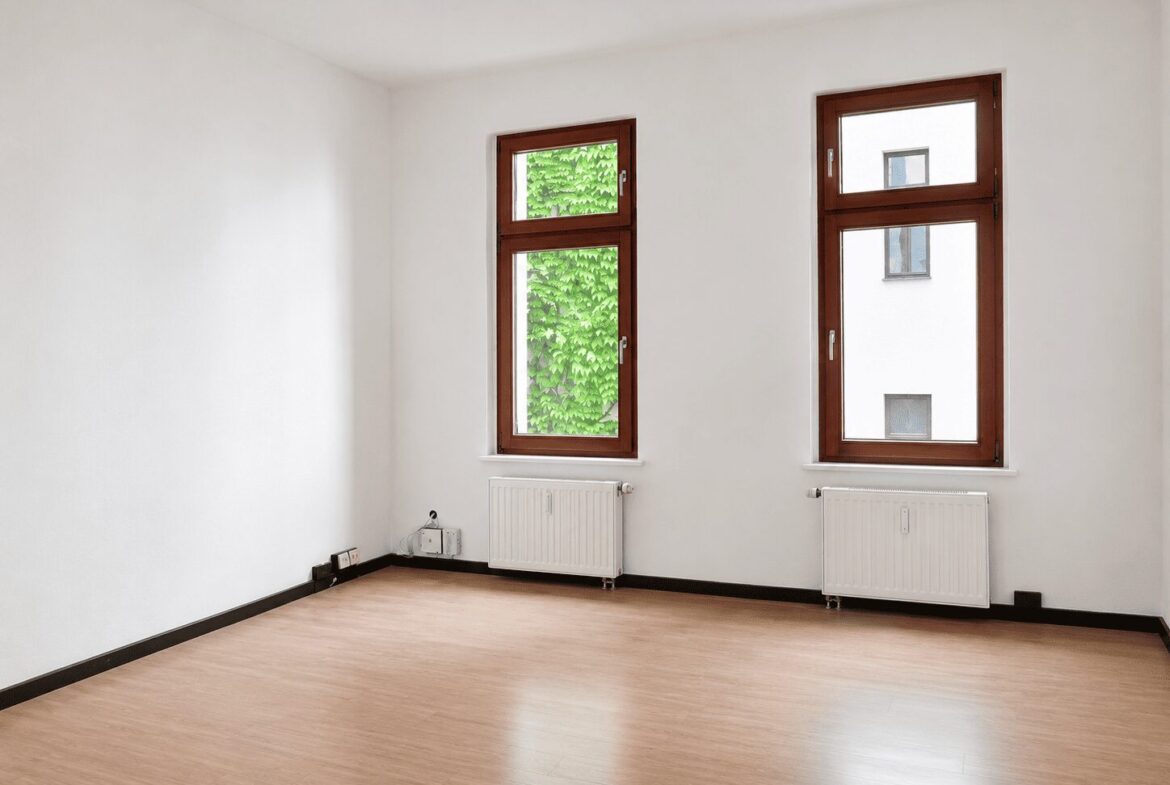 Empty white room with two tall wooden-framed windows, radiators below, and light wood flooring outside view shows greenery and a white building.