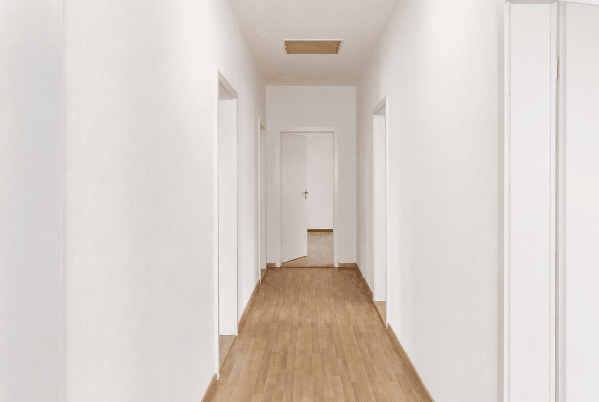 White, empty hallway with multiple doorways along the sides and a closed door at the far end, light wood flooring.