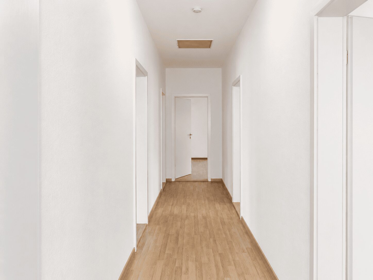 White, empty hallway with multiple doorways along the sides and a closed door at the far end, light wood flooring.