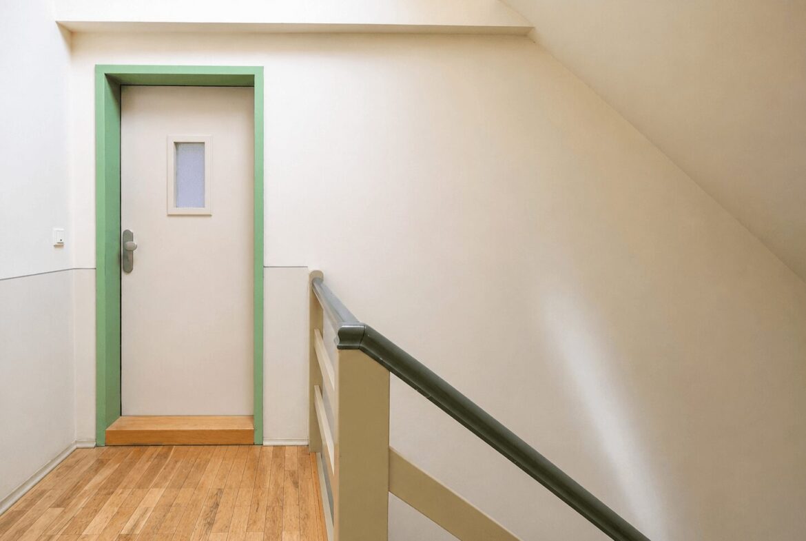 Cream-colored hallway with a lime-green-framed door at the end and a small window in the door; light wood floor and a stair railing on the right.