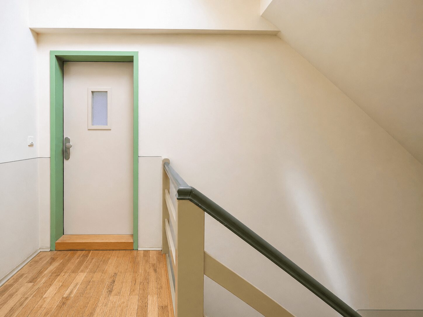 Cream-colored hallway with a lime-green-framed door at the end and a small window in the door; light wood floor and a stair railing on the right.