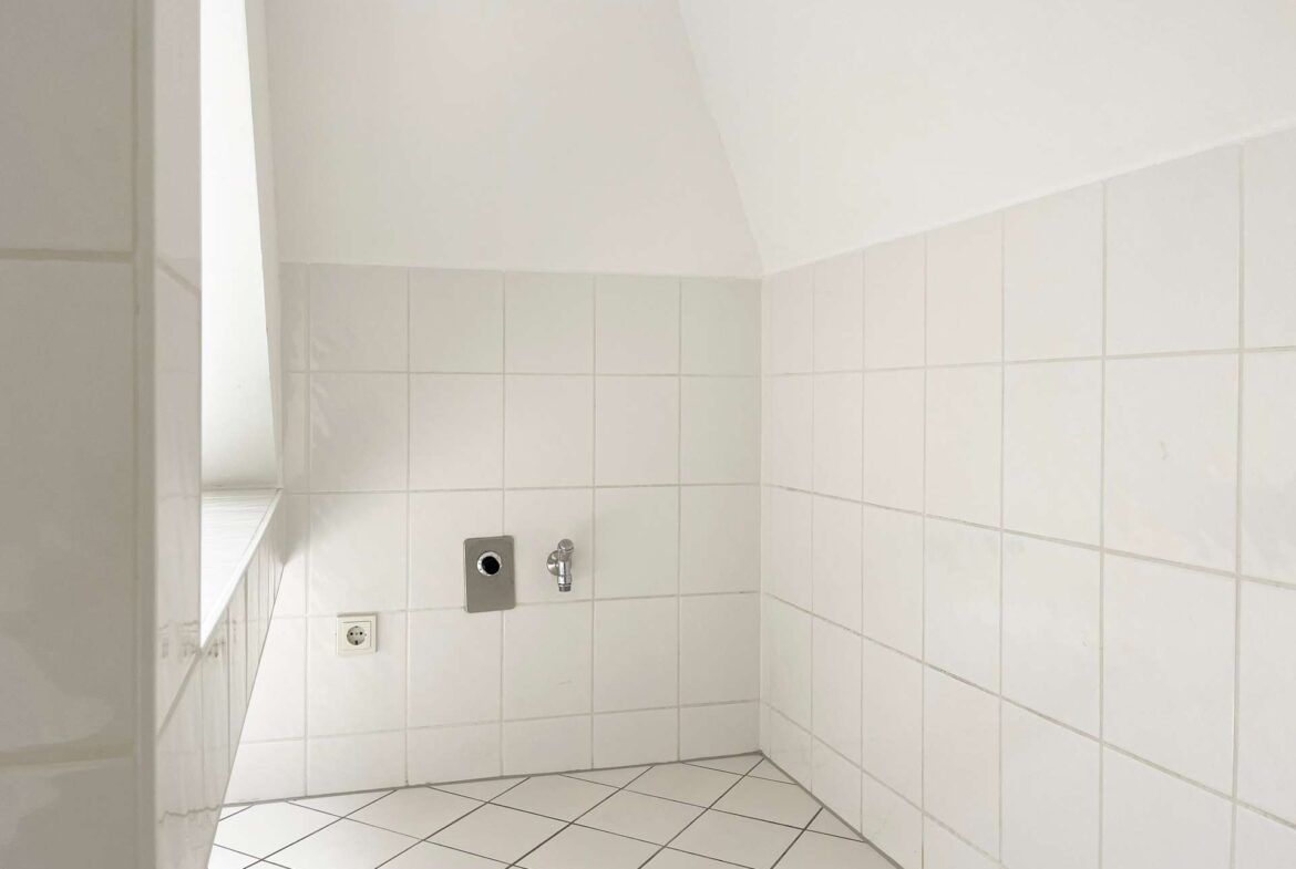 White tiled bathroom corner with a wall-mounted faucet, control plate, and a small window to the left.