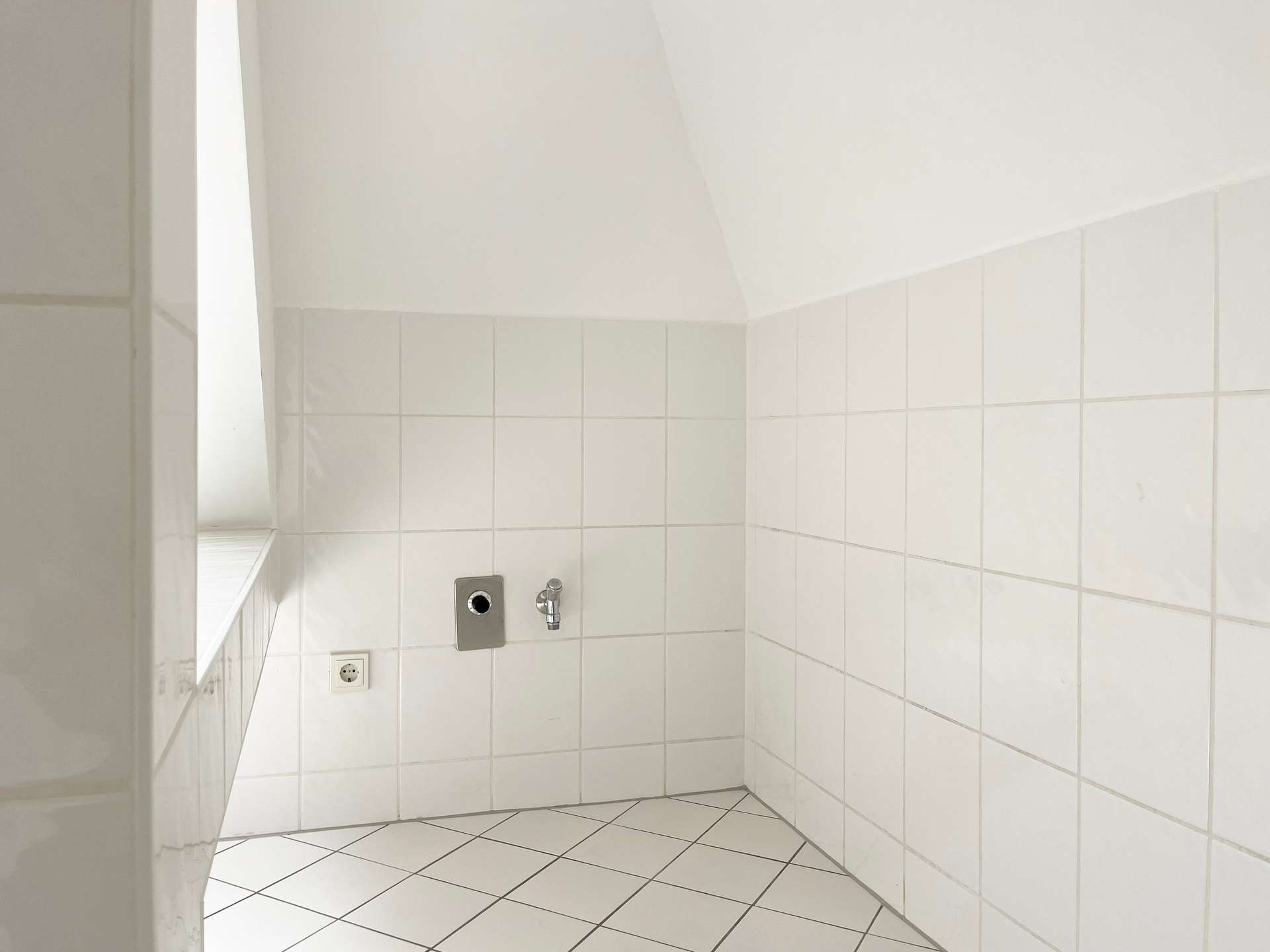 White tiled bathroom corner with a wall-mounted faucet, control plate, and a small window to the left.