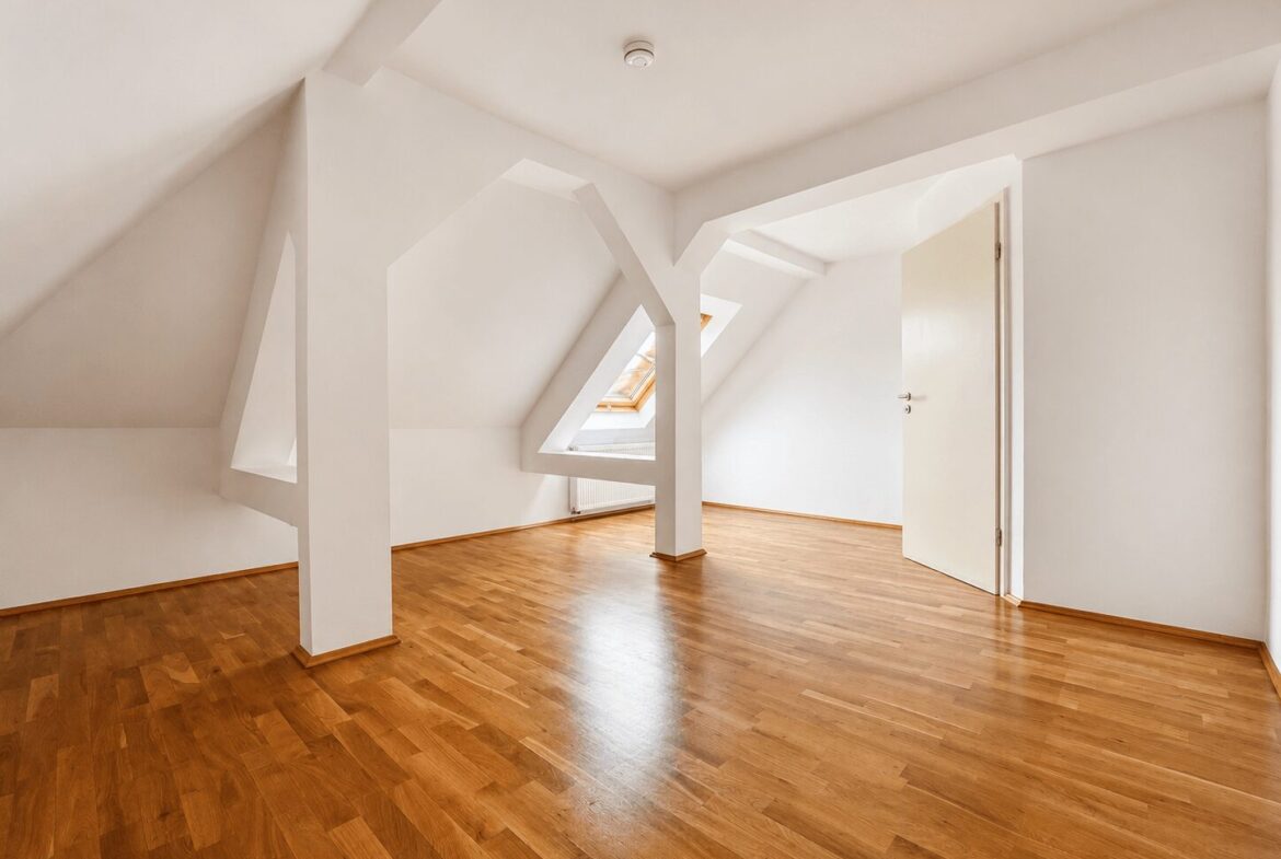 Empty attic room with white walls, wooden parquet floor, and angled sloped ceilings with triangular skylight openings.