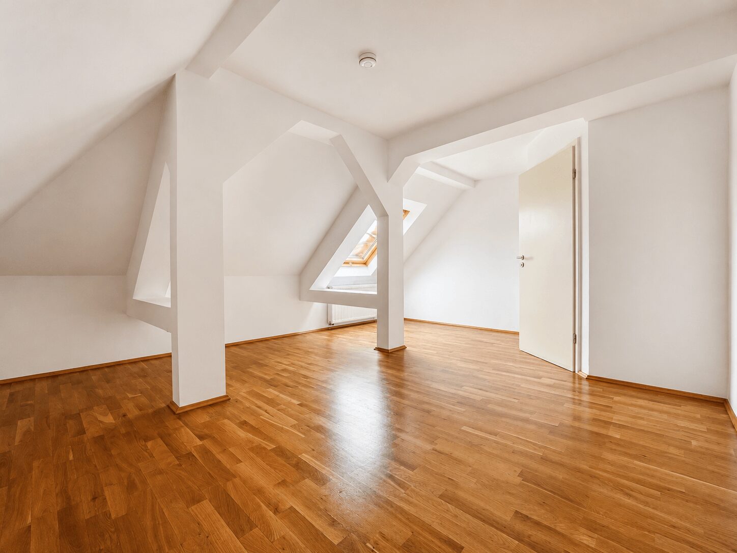 Empty attic room with white walls, wooden parquet floor, and angled sloped ceilings with triangular skylight openings.