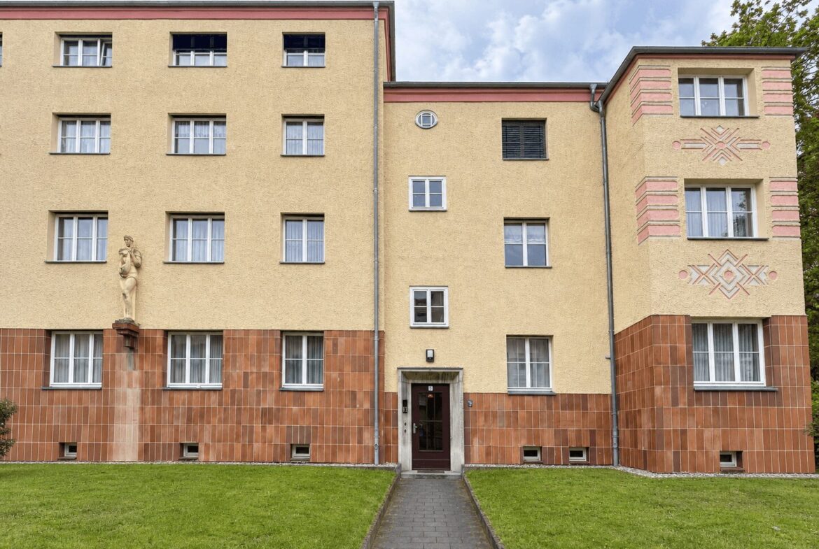 Facade of a beige residential building with a red-tiled lower wall and a central doorway, flanked by windows and a statue on the left