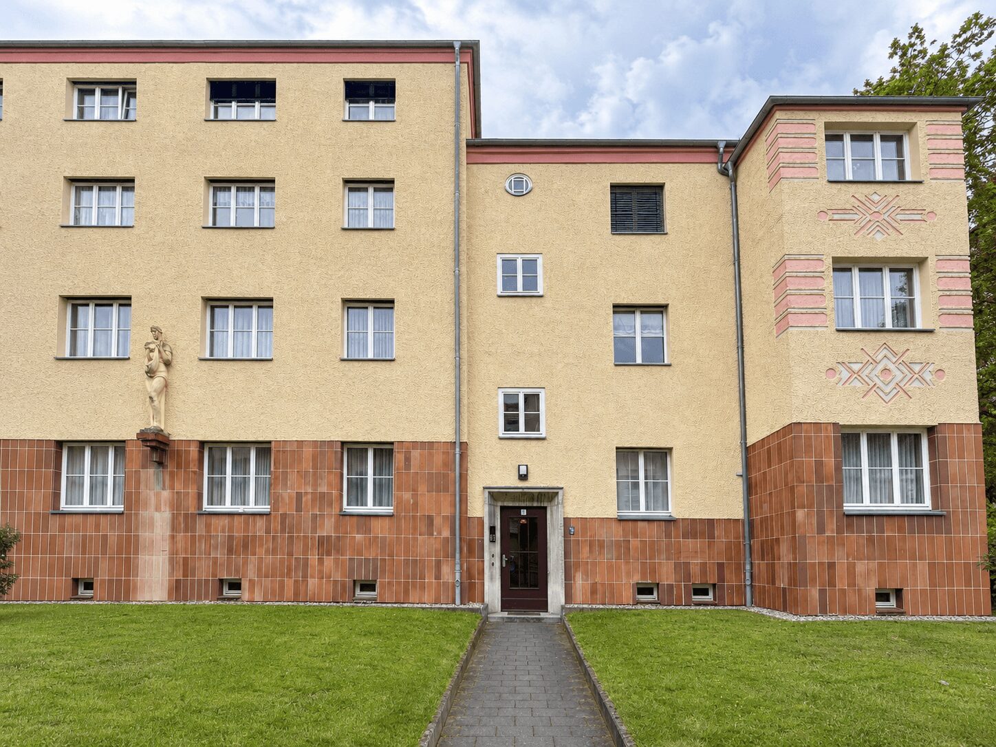 Facade of a beige residential building with a red-tiled lower wall and a central doorway, flanked by windows and a statue on the left