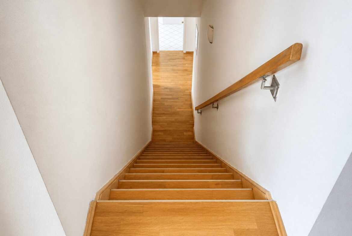 Top-down view of a wooden staircase descending to a bright hallway with a right-side handrail and white walls.