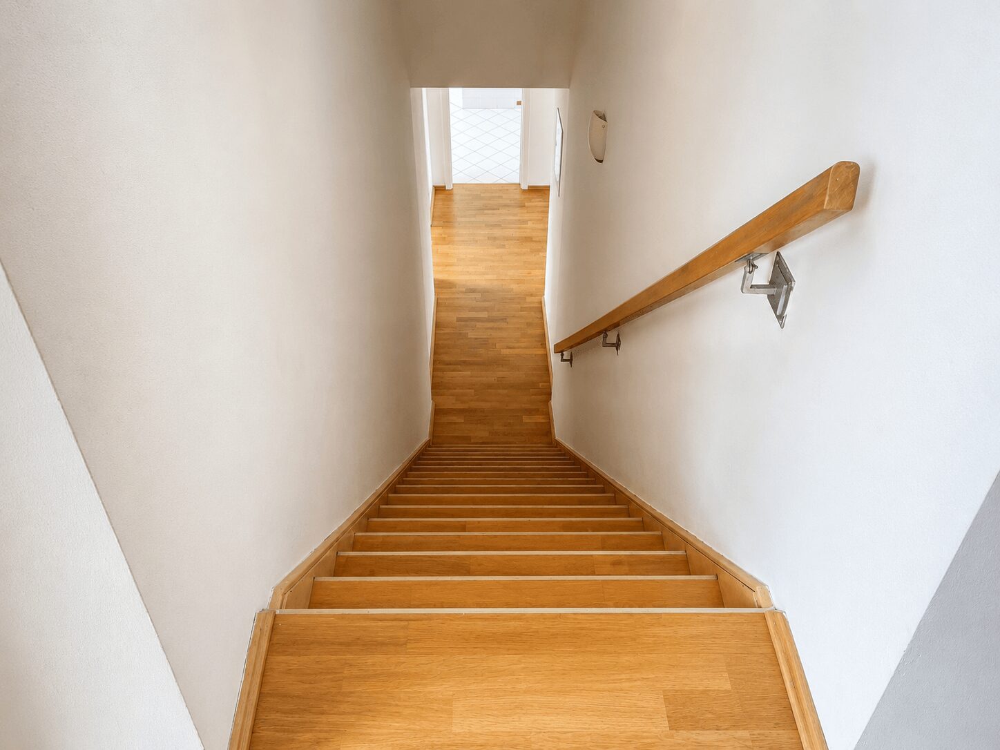 Top-down view of a wooden staircase descending to a bright hallway with a right-side handrail and white walls.